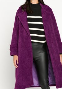 Lila texturerad kappa med stora slag, framfickor och långa ärmar, bärs över en svart-vit randig tröja och svarta byxor.