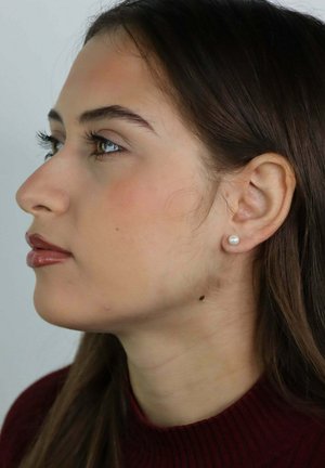 Jeune femme de profil aux cheveux bruns, portant une boucle d'oreille en perle, un haut rouge foncé et un maquillage naturel sur un fond uni.