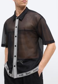 Άνδρας που φοράει μαύρο, κοντομάνικο, διάφανο πουκάμισο με κουμπιά και γκρι περιθώριο, και μαύρο σορτς με ζώνη μέσης με το λογότυπο "HUGO".