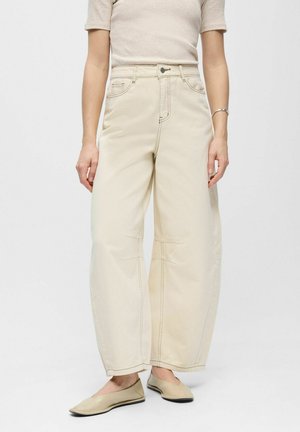 Vrouw draagt crème kleurige wijde broek met hoge taille, beige top met korte mouwen, beige platte schoenen en een zilveren armband om de linkerpols.