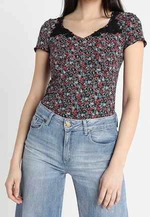 Femme portant un haut à manches courtes noir à fleurs avec une bordure en dentelle et un jean taille haute bleu clair, debout devant un fond uni.