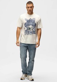 Creme t-shirt med bl�åt grafisk print med teksten "WORLD CHAMPION FOR THE SHOE SUPER BOWL XL" kombineret med lyseblå jeans og hvide sneakers.