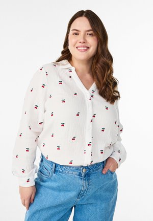 Usmievavá žena s dlhými hnedými vlasmi, oblečená v bielej košeli s červenými potlačami čerešní a modrých džínsach, s rukami v vreckách.