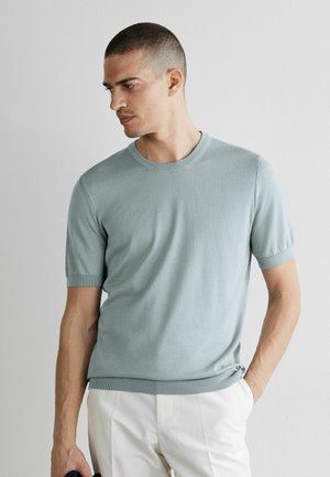 Mann mit kurzen Haaren, der einen hellblauen Kurzarm-Pullover und weiße Hosen trägt, blickt nach links, eine Hand in der Tasche.