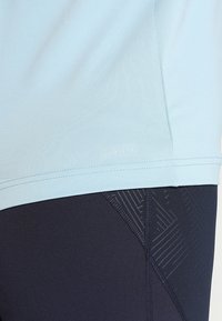 Helles blaue Sportshirt mit glatter Textur und bedrucktem Branding, kombiniert mit dunklen Leggings mit geometrischer Textur und Design.