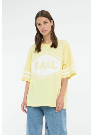 Žuta prevelika majica s bijelim heksagonalnim logotipom na kojem piše "BALL" i s dvije bijele pruge na svakom rukavu, uparena s plavim trapericama.