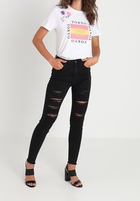Jeans skinnies rotos negros combinados con una camiseta gráfica blanca que presenta un texto colorido y acentos florales. La modelo lleva sandalias de tacón negras.
