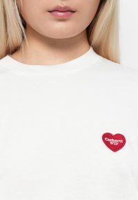Valkoinen puuvillapaita, jossa on pyöreä kaula-aukko ja lyhyet hihat. Paidassa on punainen sydämenmuotoinen brodeerattu merkki, jossa lukee "Carhartt WIP."