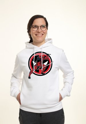 Smilende person med briller og en hvid hættetrøje med et rødt og sort Deadpool-motiv, der holder sværd, hænderne i lommerne.