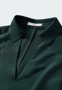 Blouse verte à col V, fabriquée en tissu lisse. Présente une patte de boutonnage et des bords bien finis. Étiquette de marque visible.