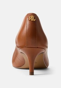 Rjav usnjen čevelj z visokim petom v zoženem oblikovanju, z zlatim logotipom na zadnji strani in tankim dizajnom pete. Gladek lesk, minimalno šivanje.