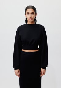 Svart cropped genser med lange ermer og rund hals, kombinert med et matchende plisséskjørt med høy midje. Glatt tekstur, minimalisk design.