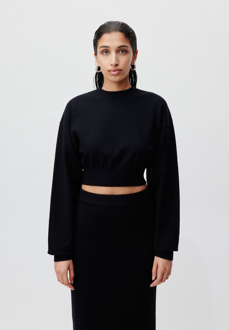 Svart cropped genser med lange ermer og rund hals, kombinert med et matchende plisséskjørt med høy midje. Glatt tekstur, minimalisk design.
