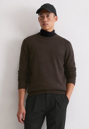 Brązowy sweter wełniany z prążkowanymi mankietami i dołem, noszony na czarnym golfie. Model ma również na sobie czapkę w kolorze czarnym oraz ciemne spodnie.