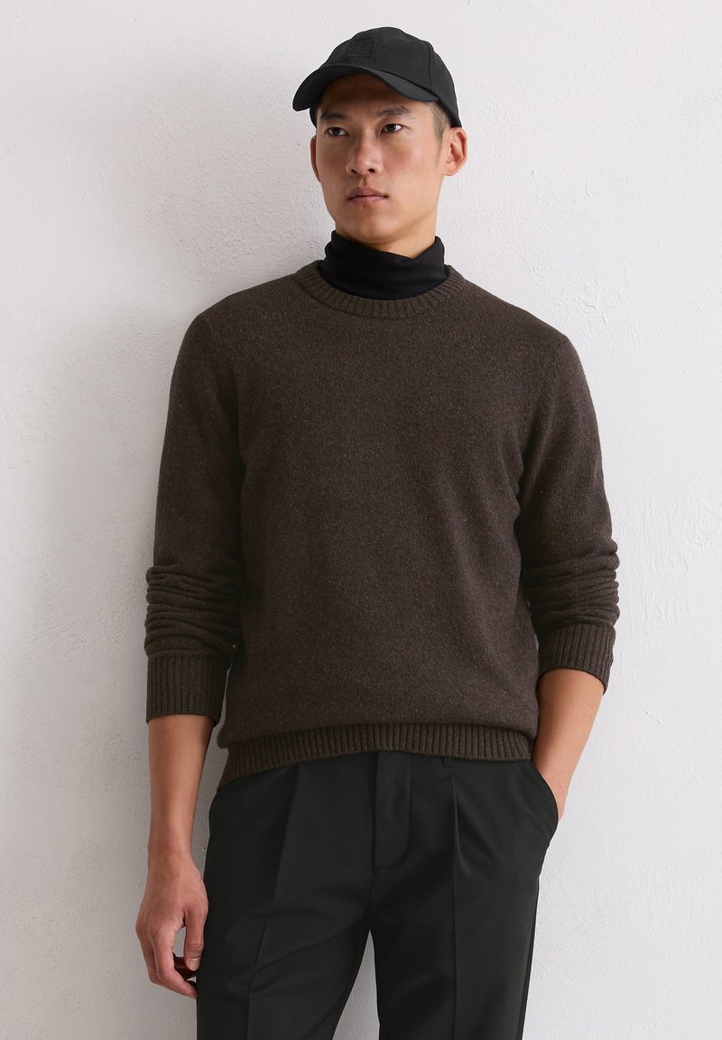 Brązowy sweter wełniany z prążkowanymi mankietami i dołem, noszony na czarnym golfie. Model ma również na sobie czapkę w kolorze czarnym oraz ciemne spodnie.