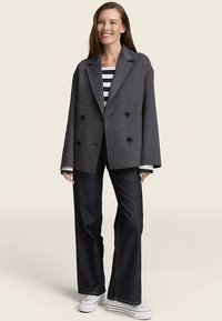 Manteau gris en laine à double boutonnage avec des boutons noirs, associé à une chemise rayée et un jean foncé. Coupe décontractée avec des manches larges.