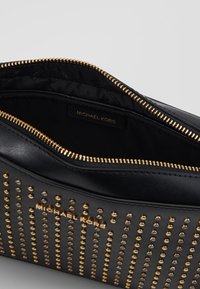 Pochette nera Michael Kors con borchie dorate disposte in linee verticali, cerniera dorata parzialmente aperta che mostra l'interno foderato con marchio.