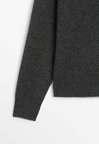 Manche et bas du torse d'un pull en maille gris foncé avec des poignets et une taille côtelés sur fond blanc.