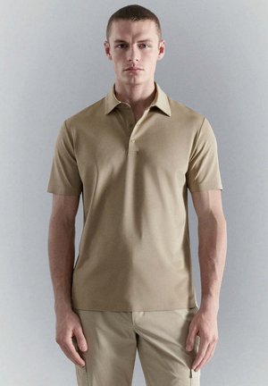 Jeune homme debout, face à l’avant, portant un polo beige à manches courtes et un pantalon beige assorti sur un fond gris uni.