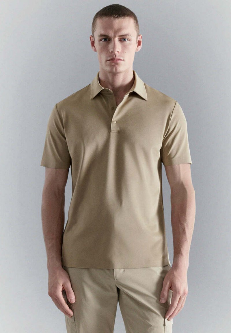 Jeune homme debout, face à l’avant, portant un polo beige à manches courtes et un pantalon beige assorti sur un fond gris uni.