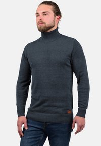 Dunkelgrauer Rollkragenpullover aus weichem Strickmaterial, mit langen Ärmeln und einem gerippten Saum sowie einem Leder-Logo-Patch an der Seite.