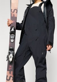 Μαύρες σκι ολόσωμες φόρμες με τσέπες με φερμουάρ και ρυθμιζόμενους ιμάντες, συνδυασμένες με σκι σε ροζ και μαύρη χρωματική παλέτα και με δέστρες.