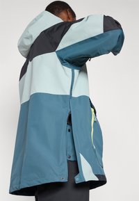 Corta-vento bicolor em tons de azul-água, preto e azul claro. Feito de material à prova de água; inclui capuz e bolsos com fecho éclair.