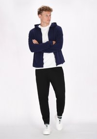 Felpa con zip di colore navy sopra una t-shirt bianca, abbinata a pantaloni jogger neri e sneakers bianche. Concentrati sul comfort atletico e sullo stile informale.