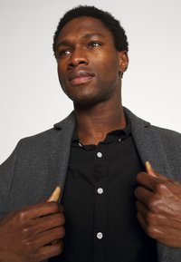 Gray textured blazer over a black button-up shirt, featuring a notch lapel and subtle stitching details, worn with hands grasping the collar.