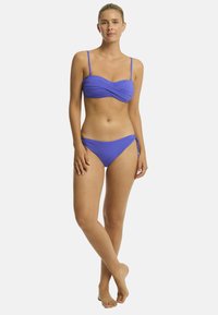 Modell trägt einen blauen Bikini, steht barfuß mit neutralem Gesichtsausdruck und präsentiert ein Badeanzugdesign vor einem schlichten weißen Hintergrund.