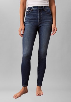 Persoon die donkere, hooggesneden skinny jeans draagt en een witte, mouwloze top, staand op blote voeten tegen een effen grijze achtergrond.