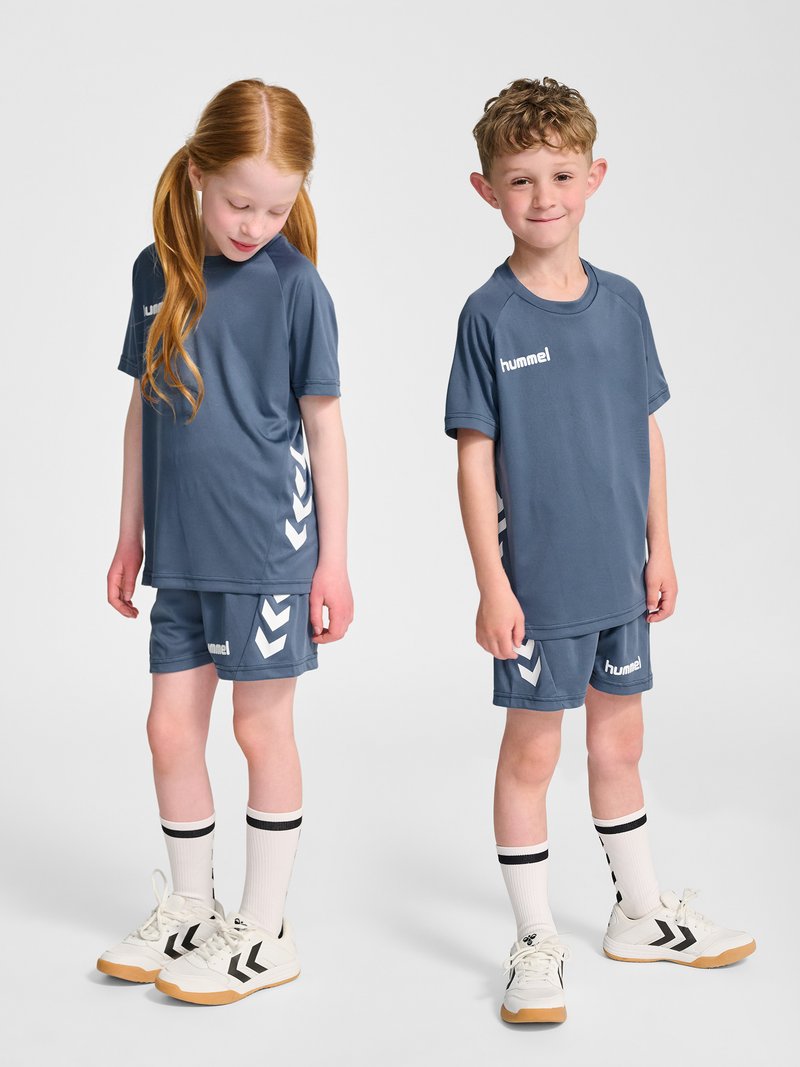 Twee kinderen in bijpassende blauwe sportkleding, witte sokken en sportschoenen staan voor een effen lichte achtergrond.