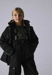 Schwarzes Winter-Außenset, bestehend aus einer dicken Jacke mit Kapuze und einem gemusterten Oberteil, kombiniert mit schwarzen Latzhosen und Cargo-Taschen.