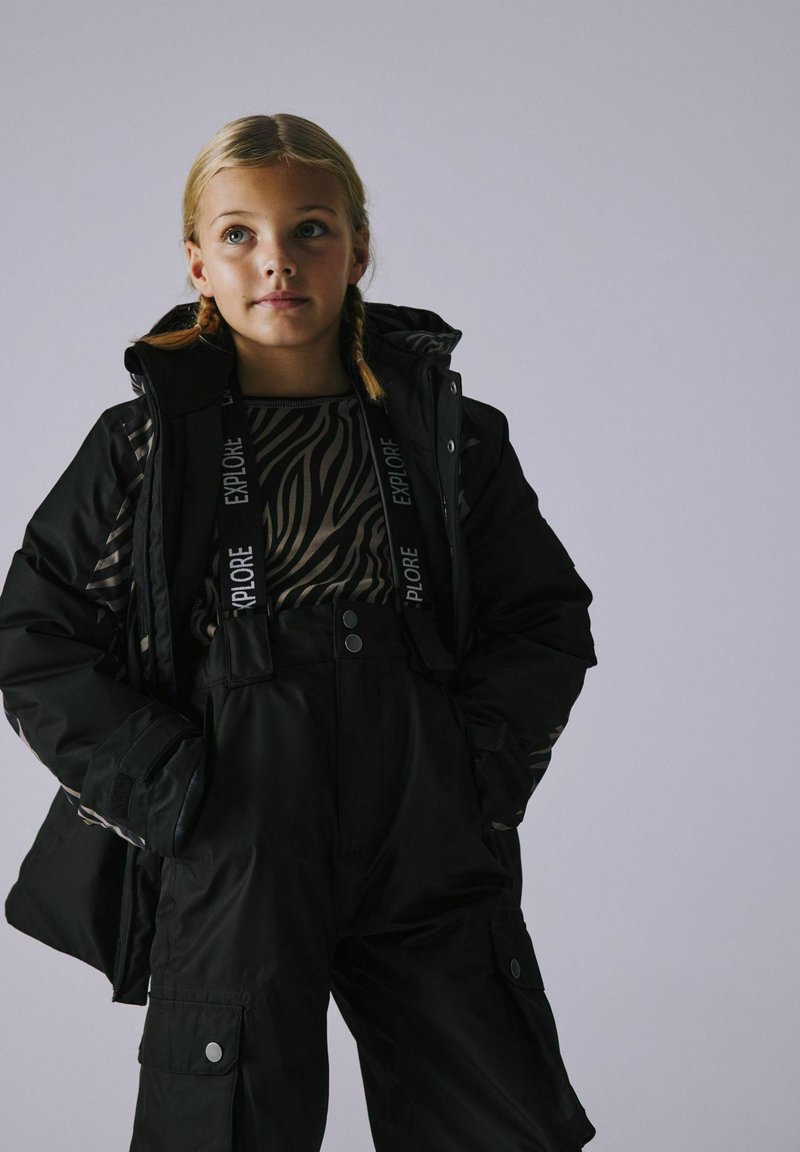 Schwarzes Winter-Außenset, bestehend aus einer dicken Jacke mit Kapuze und einem gemusterten Oberteil, kombiniert mit schwarzen Latzhosen und Cargo-Taschen.
