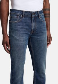 Jeans in denim di un blu scuro, con fit dritto, design a cinque tasche, cuciture arancioni a contrasto e chiusura con bottone metallico.