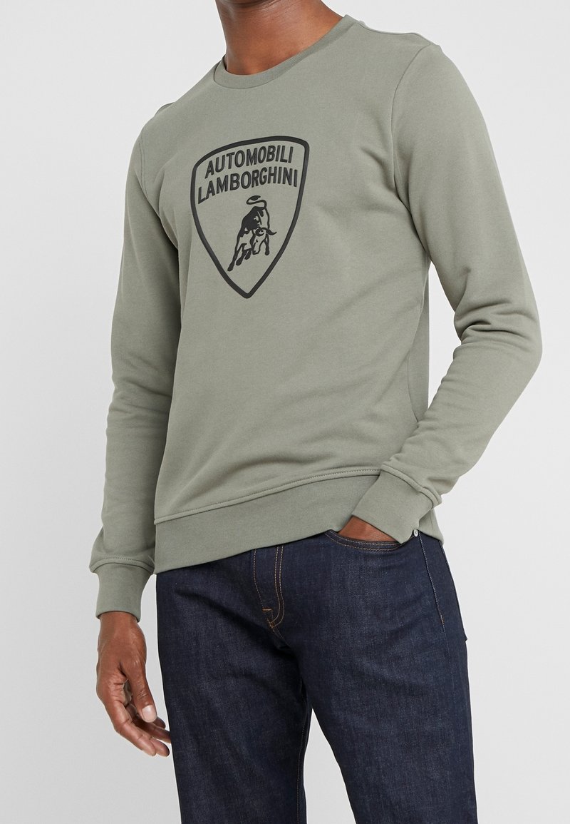 Homme portant un sweat-shirt à manches longues vert olive avec le logo noir Automobili Lamborghini et un jean bleu foncé, une main dans la poche.
