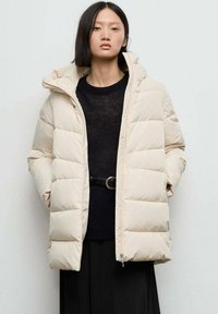 Beige pufferjack met een gewatteerd ontwerp, capuchon en ritssluiting. Gecombineerd met een zwart gebreid topje en een taille met riem, tegen een lichte achtergrond.