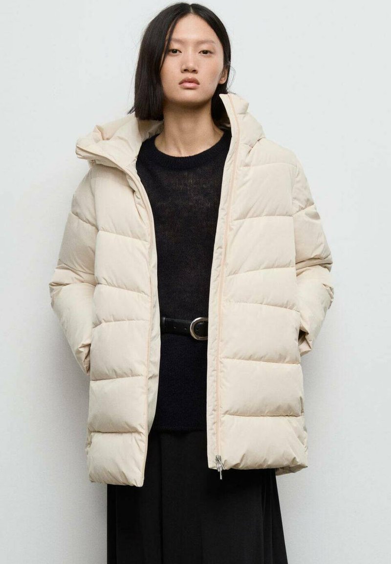 Beige pufferjack met een gewatteerd ontwerp, capuchon en ritssluiting. Gecombineerd met een zwart gebreid topje en een taille met riem, tegen een lichte achtergrond.