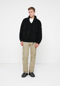 Ung man står, iförd svart zip-up fleecejacka, svart tröja, vit undertröja, beige cargobyxor och svarta sneakers mot vit bakgrund.