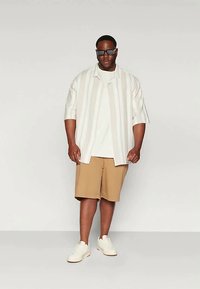 Īsām piedurknēm baltā krekls ar vertikālām svītrām, savienots ar bēšiem šortiem un baltām kedām. Modelis valkā tumšas saulesbrilles.