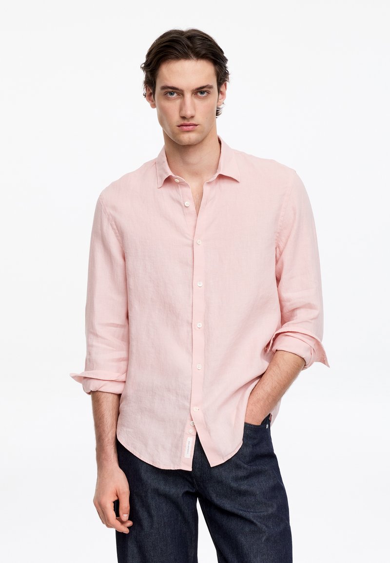 Jeune homme portant une chemise rose clair à boutons avec les manches retroussées et un jean bleu foncé, debout devant un fond blanc uni.