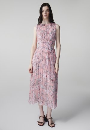 Femme portant une robe midi rose sans manches avec un imprimé corde et une découpe torsadée à la taille, associée à des sandales à talons marron, debout devant un fond gris.