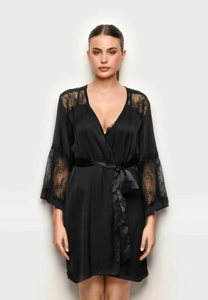 Kobieta w czarnym satynowym szlafroku z koronkowymi panelami na ramionach i rękawach, wiązanym w pasie dopasowanym paskiem, stojąca na jasnym tle.