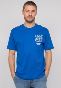 T-shirt en coton bleu à manches courtes, col rond, avec un design graphique multicolore sur la poitrine. Ourlet plat, coupe décontractée.