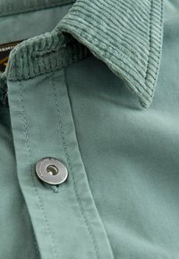 Helles grünes Hemd mit einem Cordkragen, metallischem Druckknopf und strukturiertem Stoff mit sichtbaren Steppnähten.