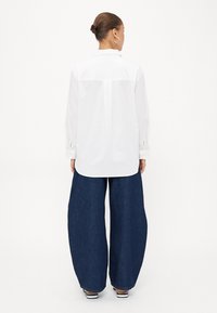 Hvit langermet skjorte med knappestolpe og et subtilt mønster, kombinert med oversized blå denimbukser. Skjorten har bakre yoke og detaljer på mansjettene.