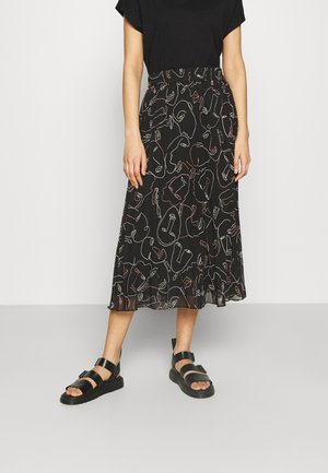Woman wearing a black midi skirt with abstract line-drawn faces, black t-shirt, and black strapped sandals standing on gray floor.