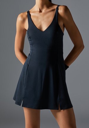 Frau trägt ein ärmelloses schwarzes Kleid mit V-Ausschnitt und seitlichen Schlitzen und steht mit den Händen hinter dem Rücken vor einem grauen Hintergrund.