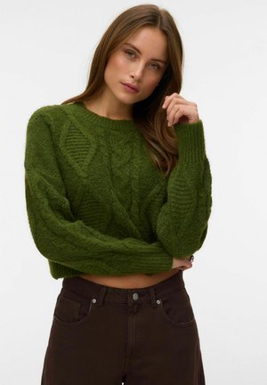 Femme aux longs cheveux bruns, portant un pull en maille torsadée vert et un pantalon foncé, posant avec une main près du visage sur un fond blanc.