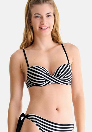 Jeune femme portant un bikini rayé noir et blanc avec un haut torsadé et un bas à nouer sur le côté, souriante sur un fond uni.